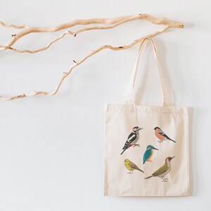 Puede incluir: Una bolsa de lona blanca con un dise&ntilde;o de cinco p&aacute;jaros diferentes. Los p&aacute;jaros son un p&aacute;jaro carpintero, un petirrojo, un mart&iacute;n pescador, un jilguero amarillo y un jilguero verde.