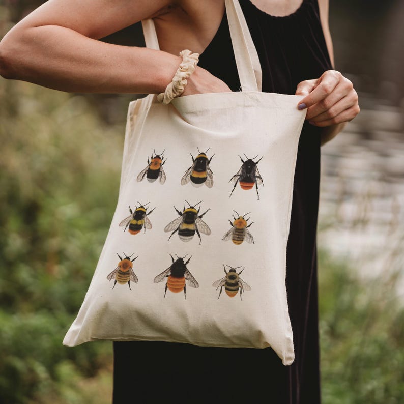 Op de afbeelding: Een witte canvas tote bag met een ontwerp van negen verschillende hommels. De bijen zijn ge&iuml;llustreerd in verschillende poses en kleuren, waaronder zwart, geel en oranje.
