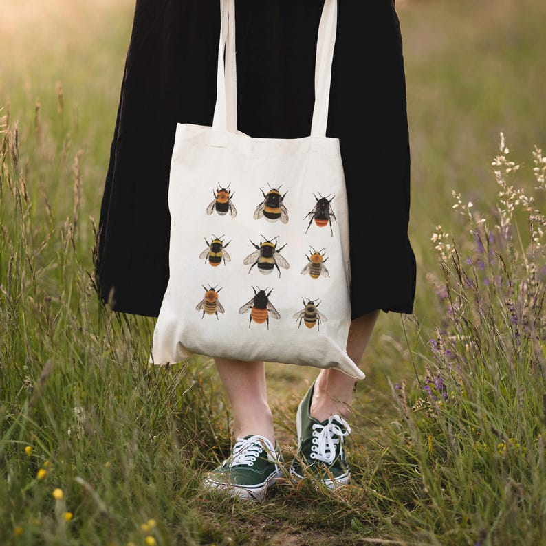 Op de afbeelding: Een witte canvas tote bag met een ontwerp van hommels in verschillende kleuren en patronen. De tas wordt gedragen door iemand die een zwarte rok en sneakers draagt.