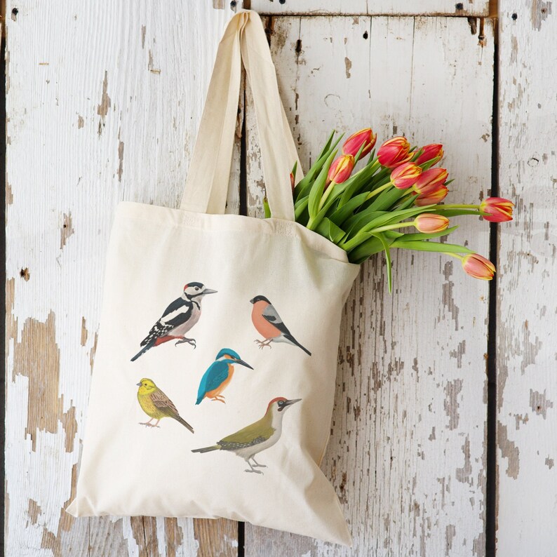 Puede incluir: Una bolsa de lona blanca con cinco ilustraciones diferentes de p&aacute;jaros. Los p&aacute;jaros son un p&aacute;jaro carpintero, un petirrojo, un mart&iacute;n pescador, un p&aacute;jaro amarillo y un p&aacute;jaro verde. La bolsa tiene un asa larga y est&aacute; hecha de tela natural.