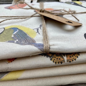 Op de afbeelding: Een stapel van vier natuurlijke canvas tassen met verschillende vogel- en vlinderontwerpen. De tassen zijn aan elkaar gebonden met touw en een bruin papieren label.