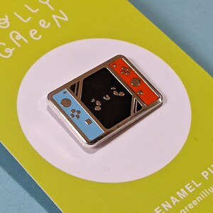 May include: A silver enamel pin featuring a stylized Nintendo Switch console with blue and red joy-cons. The pin is on a green background with the text "Holly Green" in white.