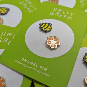 May include: Two enamel pins featuring a bee and a flower. The bee is yellow and black with a white stripe. The flower is orange with a yellow center. The pins are on a green card with the text "HOLLY GREEN" and "ENAMEL PIN" @hollygreenillustration.