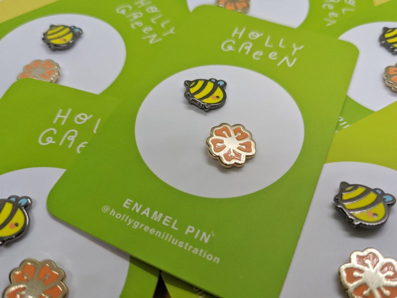 May include: Two enamel pins featuring a bee and a flower. The bee is yellow and black with a white stripe. The flower is orange with a yellow center. The pins are on a green card with the text "HOLLY GREEN" and "ENAMEL PIN" @hollygreenillustration.