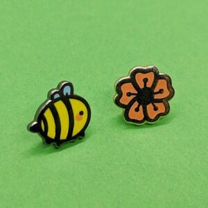 May include: Two enamel pins, one featuring a cartoon bee with black stripes and yellow body, the other a stylized orange flower with a black center.