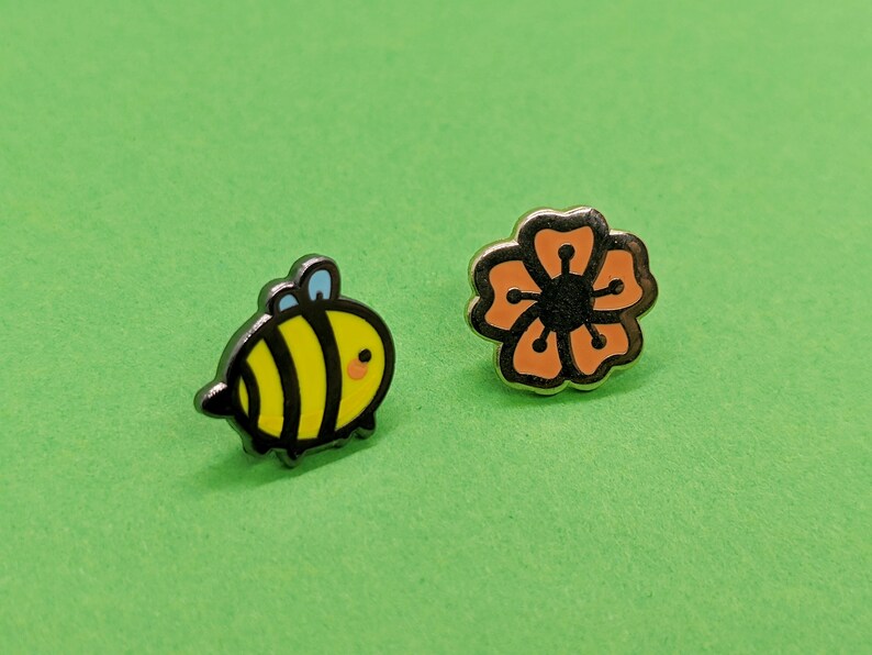 May include: Two enamel pins, one featuring a cartoon bee with black stripes and yellow body, the other a stylized orange flower with a black center.