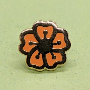 May include: A gold-colored pin with a stylized orange flower design. The flower has five petals and a black center.