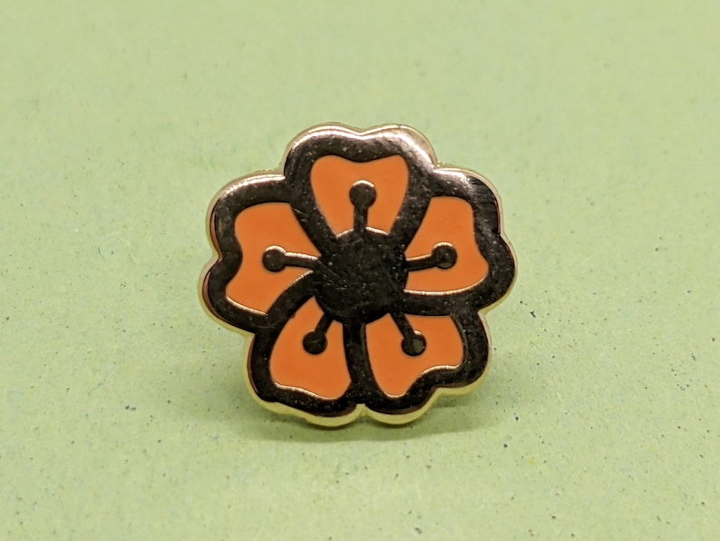 May include: A gold-colored pin with a stylized orange flower design. The flower has five petals and a black center.