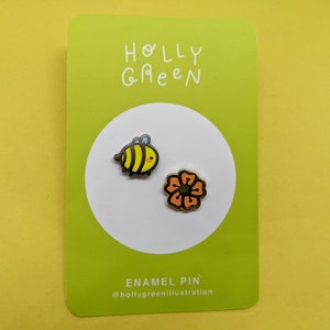 May include: A green card with the text "HOLLY GREEN" and the text "ENAMEL PIN" with the text "@hollygreenillustration". Two enamel pins are shown on the card: a yellow and black bee and an orange flower.
