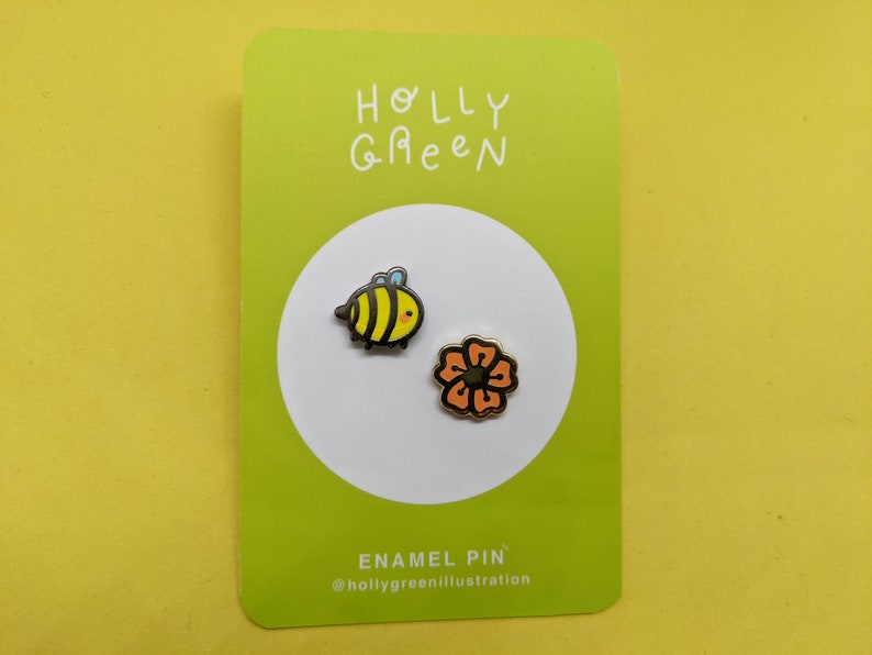 May include: A green card with the text "HOLLY GREEN" and the text "ENAMEL PIN" with the text "@hollygreenillustration". Two enamel pins are shown on the card: a yellow and black bee and an orange flower.