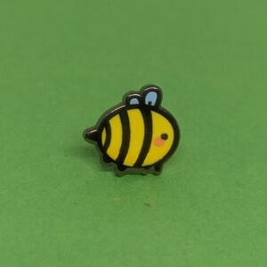 May include: A small, black and yellow enamel pin shaped like a cartoon bee with a blue antenna. The bee has a black outline and a small, pink smile.