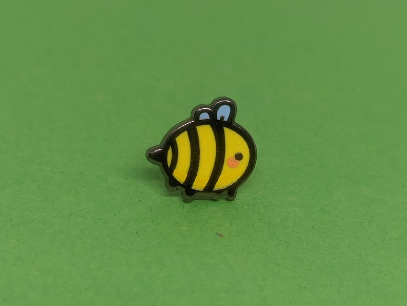 May include: A small, black and yellow enamel pin shaped like a cartoon bee with a blue antenna. The bee has a black outline and a small, pink smile.