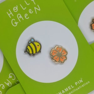 May include: Two enamel pins, one featuring a yellow and black bee with blue accents and the other a bright orange flower. The pins are displayed on a green card with the text "Holly Green Enamel Pin" and "@nillllustration".