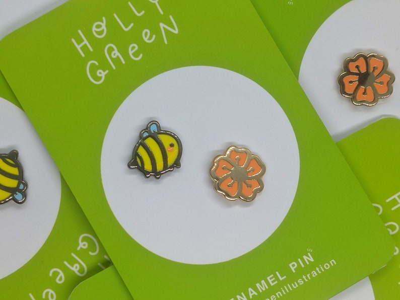 May include: Two enamel pins, one featuring a yellow and black bee with blue accents and the other a bright orange flower. The pins are displayed on a green card with the text "Holly Green Enamel Pin" and "@nillllustration".