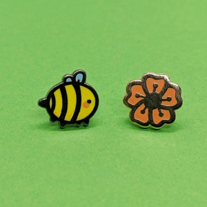 May include: Two enamel pins, one featuring a cartoon bee with black stripes and yellow body, and the other a flower with orange petals and a black center.