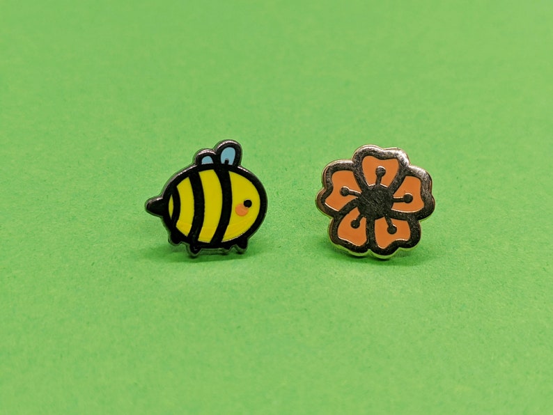 May include: Two enamel pins, one featuring a cartoon bee with black stripes and yellow body, and the other a flower with orange petals and a black center.
