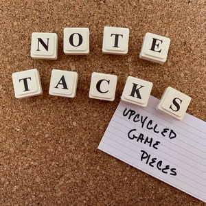 May include: White letter tiles spelling out "NOTE TACKS" on a corkboard. A white note with black handwriting reads "UPCYCLED GAME PIECES".