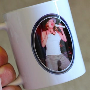 Può includere: Tazza in ceramica bianca con una foto di una donna che canta in un microfono. La foto è in una cornice circolare.