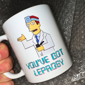 Może przedstawiać: Biały kubek ceramiczny z kreskówkowym obrazkiem lekarza ze stetoskopem i tekstem "You've got leprosy" w kolorze niebieskim i czerwonym.