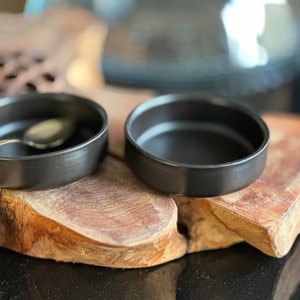 Może przedstawiać: Dwie czarne ceramiczne miski na drewnianej tacy. Miski są okrągłe i mają gładkie, błyszczące wykończenie.