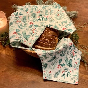 May include: A loaf of sliced bread in a woven basket, covered by a white cloth with a green and red botanical pattern. The cloth features illustrations of pine needles, holly, and berries. The scene is set on a wooden surface.