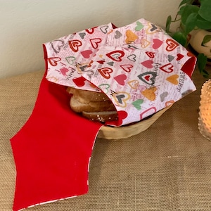 May include: A red and white fabric bread bag with a heart pattern and the words "Sweet Dreams" printed on it. The bag is filled with slices of bread and is sitting in a wooden bowl.
