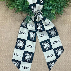 May include: A black and white scarf with a checkered pattern featuring the word "ARMY" and a military crest. The scarf is made of a soft fabric and is displayed against a backdrop of greenery and burlap.
