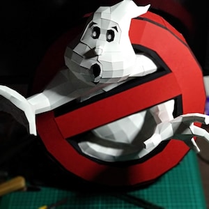 May include: A handmade papercraft of the Ghostbusters logo with the iconic ghost figure. The logo is a red circle with a white ghost inside and a red slash through it. The ghost is white with black eyes and a wide open mouth.