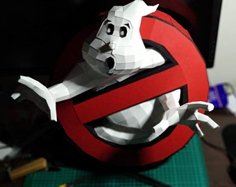 May include: A handmade papercraft of the Ghostbusters logo with the iconic ghost figure. The logo is a red circle with a white ghost inside and a red slash through it. The ghost is white with black eyes and a wide open mouth.