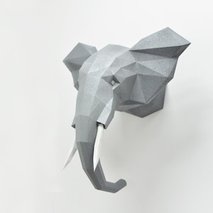 May include: A gray, geometric, 3D papercraft elephant head sculpture with white tusks. The sculpture is mounted on a white wall.