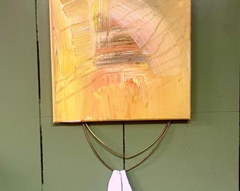 Puede incluir: Una pequeña pintura abstracta con pinceladas amarillas, naranjas y blancas. La pintura está colgada en una pared verde con un colgador de metal dorado y dos plumas de cerámica blancas colgando del colgador.