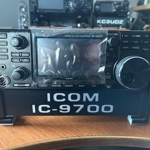 Puede incluir: Un transceptor de radioaficionado ICOM IC-9700 negro con una placa frontal plateada y una gran pantalla LCD. La radio está montada en un soporte negro con el texto "ICOM IC-9700" impreso en la parte delantera.