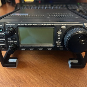 以下が含まれることがあります： 黒色のIcom IC-706MKIIG HF/VHF/UHFトランシーバー。複数のノブ、ボタン、ディスプレイ画面を備えています。ラジオは黒色のスタンドに取り付けられています。フロントパネルには「ICOM」の文字とさまざまな機能ラベルがあります。