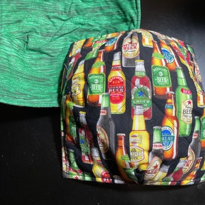 May include: Two fabric bowl cozies. One is green with a textured pattern. The other is black with a pattern of beer bottles in various colors and brands. The cozies are designed to keep bowls warm.