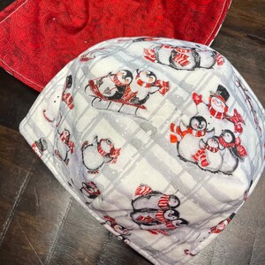 May include: A set of two red and white fabric pot holders. The smaller pot holder is red with a swirl pattern and the larger pot holder is white with a plaid pattern and penguins wearing scarves and hats.