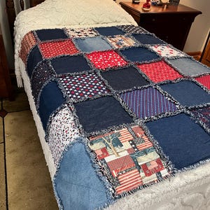May include: A handmade rag quilt featuring a patchwork design of denim squares and patriotic fabric squares with red, white, and blue star patterns, stripes, and a print containing the word Justice, all joined with exposed frayed seams.