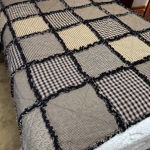 May include: A rag quilt featuring a patchwork design of square fabric blocks in black, cream, and tan. The quilt displays various patterns including gingham, stripes, and small floral prints, all joined with exposed, frayed black seams.