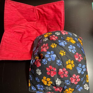 Puede incluir: Dos agarraderas de olla acolchadas. Una es roja con un patrón a rayas. La otra es negra con un patrón de huellas de patas en rojo, azul, amarillo y gris.
