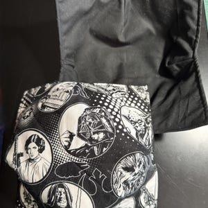 Może przedstawiać: Czarna i biała osłona na miskę z postaciami z Gwiezdnych Wojen, takimi jak Darth Vader i Yoda. Tkanina ma wzór z okręgami. Zewnętrzna strona jest czarna.
