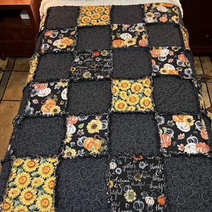 Op de afbeelding: Een patchworkquilt met vierkanten van zwarte stof met een wervelpatroon, zonnebloemen en pompoenen. De quilt heeft een witte, donzige rand en wordt op een bed getoond. De quilt heeft de woorden "gather", "Autumn", "thankful" en "happy".