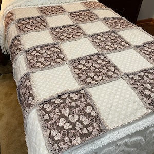 May include: A brown and white patchwork quilt with a pumpkin and leaf pattern. The quilt is made with a soft, fluffy fabric and has a frayed edge.