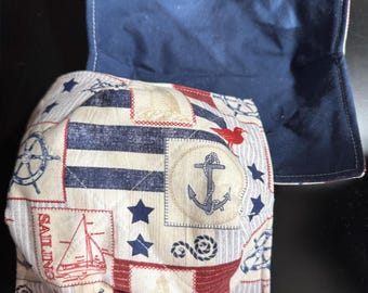 May include: Nautical-themed bowl cozy in navy blue, red, and cream. The cozy features a patchwork design with anchors, sailboats, stars, and the word "SAILING". The navy blue backing is visible.
