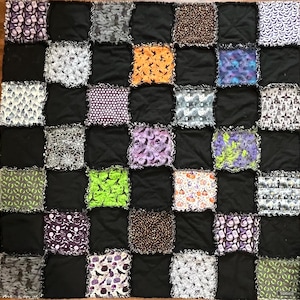 Könnte beinhalten: Ein Patchwork-Quilt mit Halloween-Thema. Der Quilt zeigt ein Raster aus schwarzen Quadraten, die mit Stoffquadraten mit Mustern von Fledermäusen, Kürbissen und anderen gruseligen Designs durchsetzt sind. Die Kanten der Quadrate sind ausgefranst.