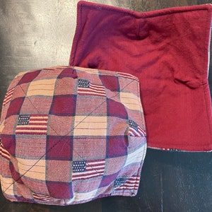Puede incluir: Dos agarraderas de olla acolchadas de color rojo y marrón con un patrón de cuadros y pequeños diseños de banderas americanas. Las agarraderas de olla están hechas de tela y tienen un diseño acolchado.