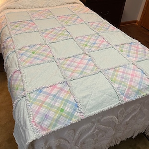 May include: A light blue and white patchwork quilt with a checkered pattern in pastel colors. The quilt is made of soft, fluffy fabric and has a fringed edge.