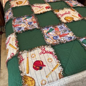 May include: A baseball-themed quilt featuring a patchwork design. The quilt combines green squares with baseball-themed fabric squares. The baseball squares display baseball bats, gloves, helmets, and baseballs. The quilt has a white, fluffy border.