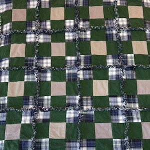 Könnte beinhalten: Eine handgefertigte Patchwork-Quilt mit einem Rastermuster aus grünen, beigen und karierten Quadraten. Der Quilt hat einen Fransenrand, der Textur verleiht. Die karierten Quadrate enthalten Blau-, Weiß- und Grüntöne.