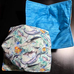 May include: Two fabric bowl cozies. One features a blue and white ocean-themed print with whales, sharks, and octopuses. The other is a solid blue with a textured appearance. These cozies are designed to hold bowls and protect hands from heat.