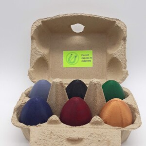 Puede incluir: Una huevera de cartón con seis huevos de madera pintados de varios colores: azul, azul oscuro, negro, rojo, verde y naranja. Una pegatina verde con texto es visible en la huevera.