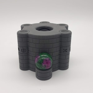 Puede incluir: Una caja de rompecabezas hexagonal gris, impresa en 3D, con una canica verde y rosa en su interior. La caja tiene un patrón de ladrillo y una abertura circular en la parte superior.